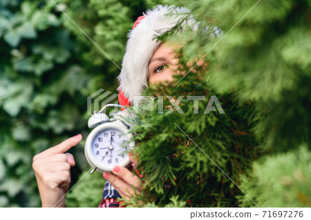 Face of woman in a santa claus red hat holding alarm clock in green fir tree branches in the forest. Christmas gift concept. Hurry up, shopping, delivery, sale Face of woman in a santa claus red hat holding alarm clock in green fir tree branches in the forest. Christmas gift concept. Hurry up, shopping, delivery, sale 71697276