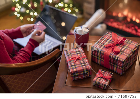 Woman holding credit card using laptop for making order sitting at table with packaging gift, cup of cocoa and marshmallows near fireplace and christmas tree. Online shopping concept Woman holding credit card using laptop for making order sitting at table with packaging gift, cup of cocoa and marshmallows near fireplace and christmas tree. Online shopping concept 71697752