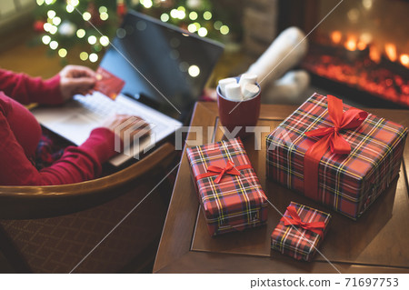 Woman holding credit card using laptop for making order sitting at table with packaging gift, cup of cocoa and marshmallows near fireplace and christmas tree. Online shopping concept Woman holding credit card using laptop for making order sitting at table with packaging gift, cup of cocoa and marshmallows near fireplace and christmas tree. Online shopping concept 71697753