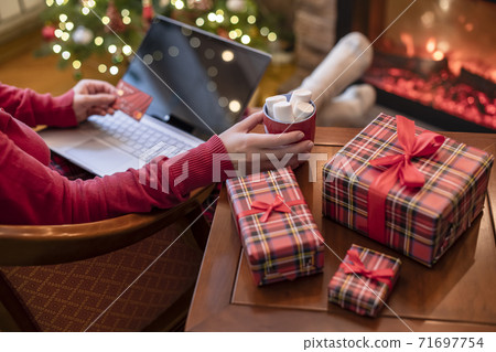 Woman holding credit card using laptop for making order sitting at table with packaging gift, cup of cocoa and marshmallows near fireplace and christmas tree. Online shopping concept Woman holding credit card using laptop for making order sitting at table with packaging gift, cup of cocoa and marshmallows near fireplace and christmas tree. Online shopping concept 71697754