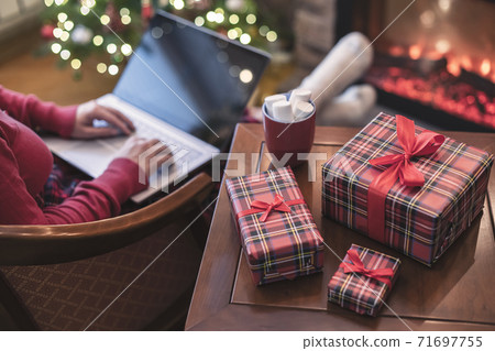 Woman holding credit card using laptop for making order sitting at table with packaging gift, cup of cocoa and marshmallows near fireplace and christmas tree. Online shopping concept Woman holding credit card using laptop for making order sitting at table with packaging gift, cup of cocoa and marshmallows near fireplace and christmas tree. Online shopping concept 71697755