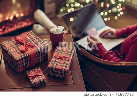 Woman holding credit card using laptop for making order sitting at table with packaging gift, cup of cocoa and marshmallows near fireplace and christmas tree. Online shopping concept Woman holding credit card using laptop for making order sitting at table with packaging gift, cup of cocoa and marshmallows near fireplace and christmas tree. Online shopping concept 71697756