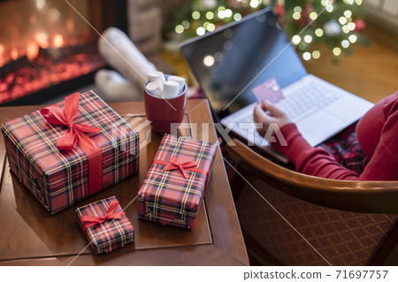 Woman holding credit card using laptop for making order sitting at table with packaging gift, cup of cocoa and marshmallows near fireplace and christmas tree. Online shopping concept 71697757