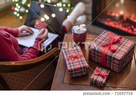 Woman holding credit card using laptop for making order sitting at table with packaging gift, cup of cocoa and marshmallows near fireplace and christmas tree. Online shopping concept Woman holding credit card using laptop for making order sitting at table with packaging gift, cup of cocoa and marshmallows near fireplace and christmas tree. Online shopping concept 71697758