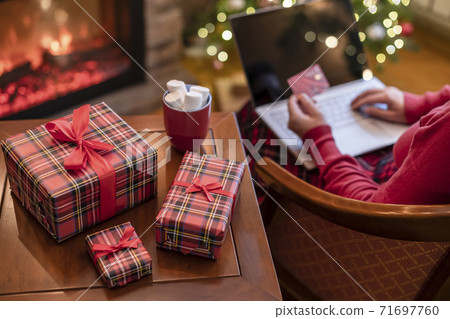 Woman holding credit card using laptop for making order sitting at table with packaging gift, cup of cocoa and marshmallows near fireplace and christmas tree. Online shopping concept Woman holding credit card using laptop for making order sitting at table with packaging gift, cup of cocoa and marshmallows near fireplace and christmas tree. Online shopping concept 71697760