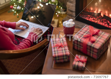 Christmas. Woman using laptop for searching gift ideas sitting at table with ready gift boxes and cup of cocoa and marshmallows near fireplace and christmas tree. Concept Christmas. Woman using laptop for searching gift ideas sitting at table with ready gift boxes and cup of cocoa and marshmallows near fireplace and christmas tree. Concept 71697767
