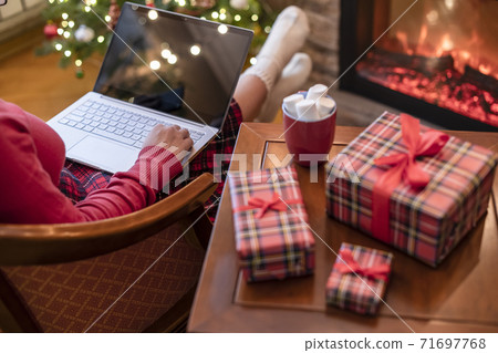 Christmas. Woman using laptop for searching gift ideas sitting at table with ready gift boxes and cup of cocoa and marshmallows near fireplace and christmas tree. Concept Christmas. Woman using laptop for searching gift ideas sitting at table with ready gift boxes and cup of cocoa and marshmallows near fireplace and christmas tree. Concept 71697768