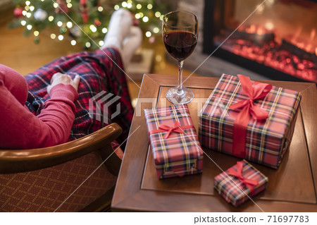 Alone woman covered plaid with glass of wine sitting and relaxation on armchair near fareplace and christmas tree after finishing pakking gift boxes for family. Alone woman covered plaid with glass of wine sitting and relaxation on armchair near fareplace and christmas tree after finishing pakking gift boxes for family. 71697783