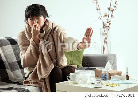 Woman wrapped in a plaid looks sick, ill, sneezing and coughing sitting at home indoors Woman wrapped in a plaid looks sick, ill, sneezing and coughing sitting at home indoors 71703991