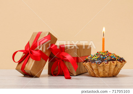 Tasty birthday muffin with burning festive candle and gift boxes on beige background. Tasty birthday muffin with burning festive candle and gift boxes on beige background. 71705103