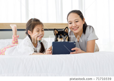 Happy Asian mother and daughter lying on bed and using digital tablet. Black Shiba Inu on bed between its owner. Concept family with dog at home. 71710339