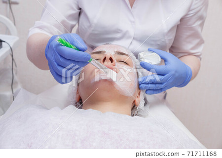 Master creates a white mask with a brush on the face of a young woman lying on the table. 71711168