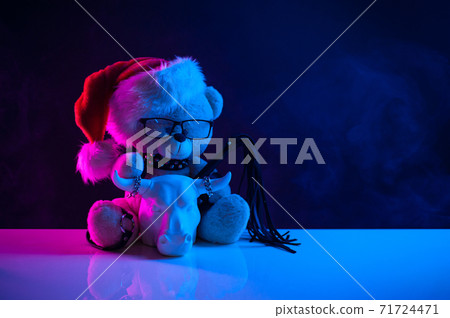 a toy bear in a Santa Claus hat wearing leather straps harness accessory for BDSM along with a statuette of a white bull on a dark background in neon light a toy bear in a Santa Claus hat wearing leather straps harness accessory for BDSM along with a statuette of a white bull on a dark background in neon light 71724471