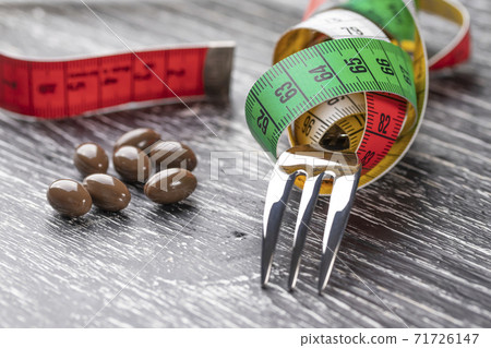 A shiny fork wrapped in a multi-colored centimeter to measure body shapes and brown medical capsules on a black old wooden table. The concept of nutritional supplements for weight loss 71726147