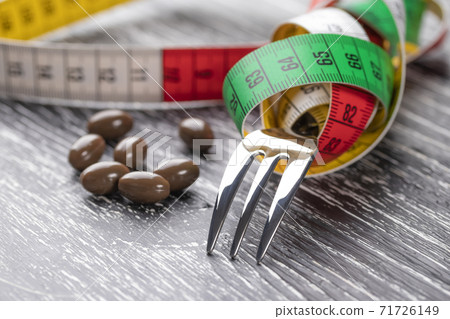 A shiny fork wrapped in a multi-colored centimeter to measure body shapes and brown medical capsules on a black old wooden table. The concept of nutritional supplements for weight loss A shiny fork wrapped in a multi-colored centimeter to measure body shapes and brown medical capsules on a black old wooden table. The concept of nutritional supplements for weight loss 71726149