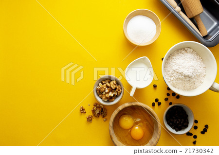 Ingredients for baking - flour, sugar, milk, eggs, wallnuts, chocolate chips, rolling pin, baking mold. Yellow background with copy space. 71730342