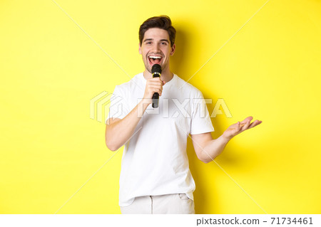 Happy man entertainer performing, talking in microphone, making speech or stand-up show, standing over yellow background Happy man entertainer performing, talking in microphone, making speech or stand-up show, standing over yellow background 71734461