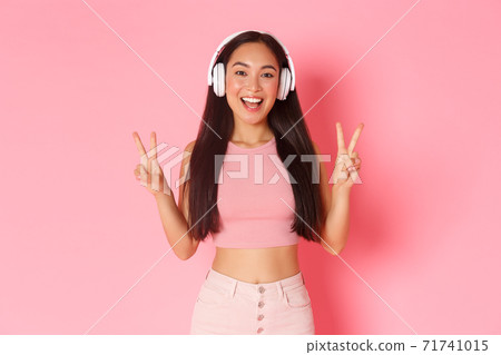 Portrait of beautiful and upbeat, smiling asian girl listening music in headphones, showing peace kawaii gesture, sending positive vibes, standing over pink background Portrait of beautiful and upbeat, smiling asian girl listening music in headphones, showing peace kawaii gesture, sending positive vibes, standing over pink background 71741015