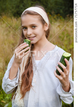1 white girl 11 years old in a white shirt eating a fresh cucumber on the background of grass 71757714