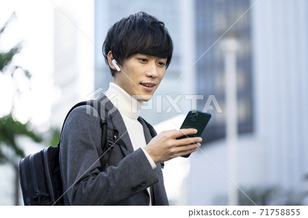 一個年輕人戴著耳機，看著他的智能手機的屏幕 71758855