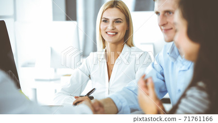 Group of business people discussing questions at meeting. Headshot of blonde businesswoman while smiling to her colleague at office negotiation. Teamwork and cooperation in corporate occupation Group of business people discussing questions at meeting. Headshot of blonde businesswoman while smiling to her colleague at office negotiation. Teamwork and cooperation in corporate occupation 71761166
