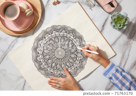 Top view of colored mandala art book with colorful markers, pen, pink pen case and a cup of coffee and macaroons. White marble background. 71763170