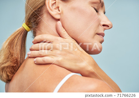 Suffering from back pain. Rear view of a mature woman touching her neck while standing against blue background Suffering from back pain. Rear view of a mature woman touching her neck while standing against blue background 71766226
