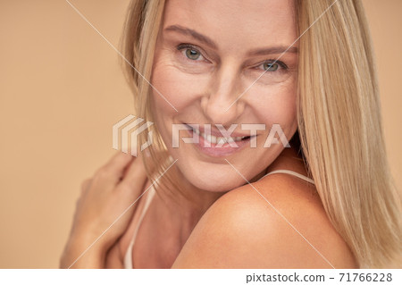 Portrait of beautiful adult woman touching her shoulders with her hands and looking at camera while posing in studio over beige background Portrait of beautiful adult woman touching her shoulders with her hands and looking at camera while posing in studio over beige background 71766228