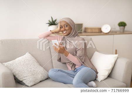 Young black woman in hijab using smartphone for work or studies on sofa at home Young black woman in hijab using smartphone for work or studies on sofa at home 71766999