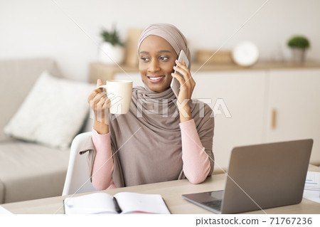 Lovely black lady in hijab talking on smartphone and drinking coffee near laptop computer at home Lovely black lady in hijab talking on smartphone and drinking coffee near laptop computer at home 71767236