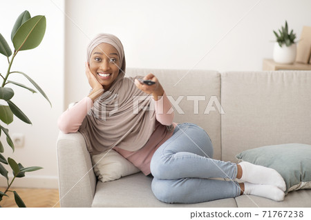 Positive black woman in hijab watching TV on couch at home, copy space 71767238