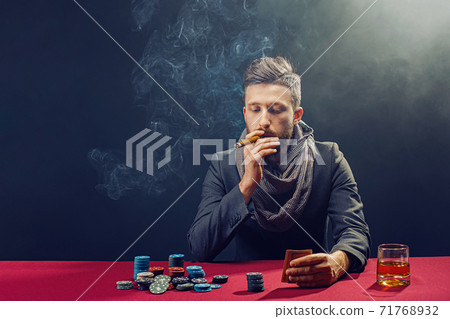 Stylish bearded Man in suit and scarf playing in dark casino, smoking cigar, drink whiskey Stylish bearded Man in suit and scarf playing in dark casino, smoking cigar, drink whiskey 71768932