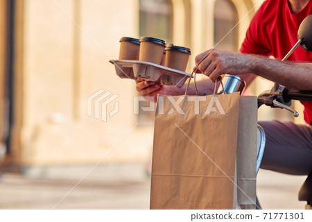 Male courier delivering four coffee cups and paper bag with food by scooter 71771301