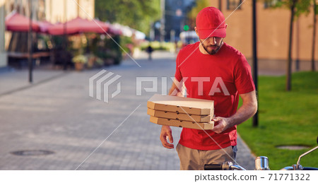Young caucasian delivery man in red uniform delivering pizza, standing near his scooter Young caucasian delivery man in red uniform delivering pizza, standing near his scooter 71771322