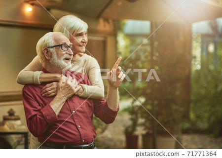 Smiling male retiree in glasses pointing finger to the side and embracing with his wife Smiling male retiree in glasses pointing finger to the side and embracing with his wife 71771364