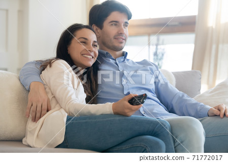 Happy young spouses cuddling on couch at home watching tv Happy young spouses cuddling on couch at home watching tv 71772017