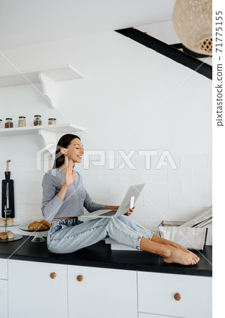 Image of cute brunette woman sitting at kitchen and using laptop 71775155