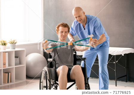 Mature clinician in blue uniform supporting hand of young patient in wheelchair Mature clinician in blue uniform supporting hand of young patient in wheelchair 71776925
