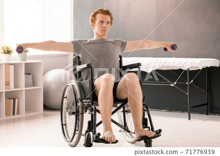 Young sportsman sitting in wheelchair and outstretching arms with dumbbells Young sportsman sitting in wheelchair and outstretching arms with dumbbells 71776939