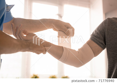 Hands of clinician wrapping elbow of right arm of sick young man with bandage Hands of clinician wrapping elbow of right arm of sick young man with bandage 71776940