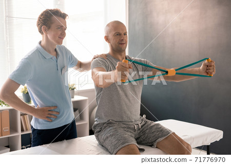 Bald man stretching resistance band during exercise for arms on medical couch 71776970