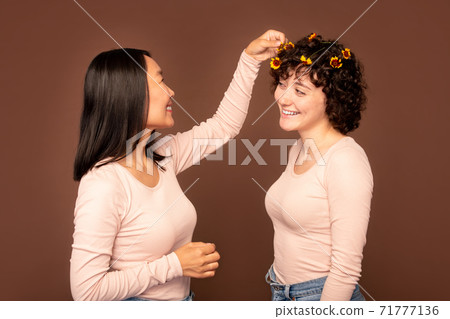 one of two young intercultural women putting small flower in hair of her friend one of two young intercultural women putting small flower in hair of her friend 71777136