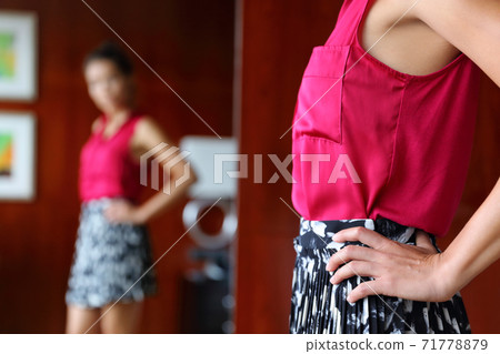 Woman getting ready in the morning trying on clothing choosing what to wear for work looking in mirror at home. Weight loss body self-esteem concept or fashion choice indecision. Woman getting ready in the morning trying on clothing choosing what to wear for work looking in mirror at home. Weight loss body self-esteem concept or fashion choice indecision. 71778879