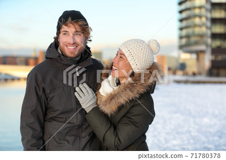 Winter couple in snow hat and coats. Happy young adults walking together smiling on a city street enjoying sunny urban walk in modern neighborhood. Asian girlfriend in love with man. 71780378