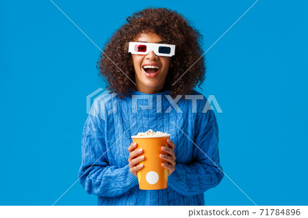 Cheerful gorgeous african-american woman enjoying watching movie in cinema, laughing and smiling excited with 3d effects wear glasses, holding popcorn, standing blue background 71784896