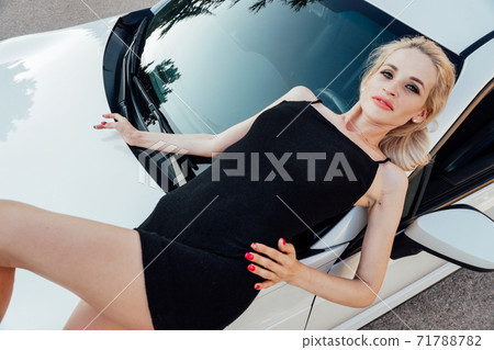 Beautiful blonde woman in black dress on the hood of sports car Beautiful blonde woman in black dress on the hood of sports car 71788782