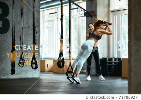 TRX workout. Young athletic woman in sportswear exercising with fitness straps with assistance of personal trainer at gym 71788927