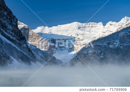 Lake Louise in early winter sunny day morning. Mist floating on turquoise color water surface. Clear blue sky, snow capped mountains in background. Beautiful natural landscape in Banff National Park. 71789906