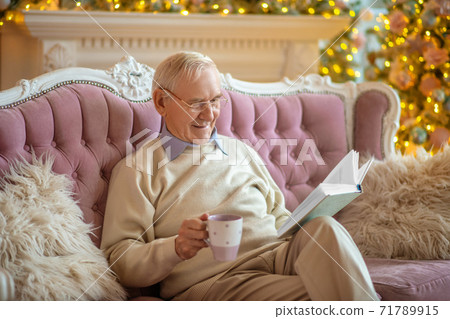 Grey-haired man sitting on a sofa and reading a book 71789915