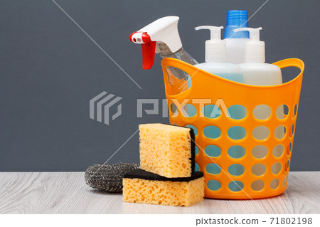 Bottles of dishwashing liquid and sponges in a basket on wooden desk. Bottles of dishwashing liquid and sponges in a basket on wooden desk. 71802198